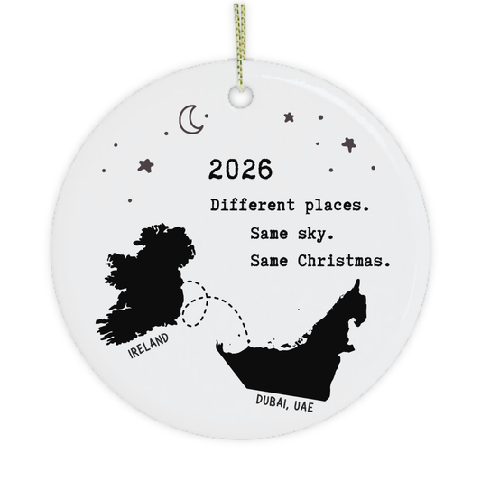 A round white ceramic Christmas ornament with a gold hanging string, featuring black printed graphics of Ireland and the United Arab Emirates with the text '2026 Different places. Same sky.