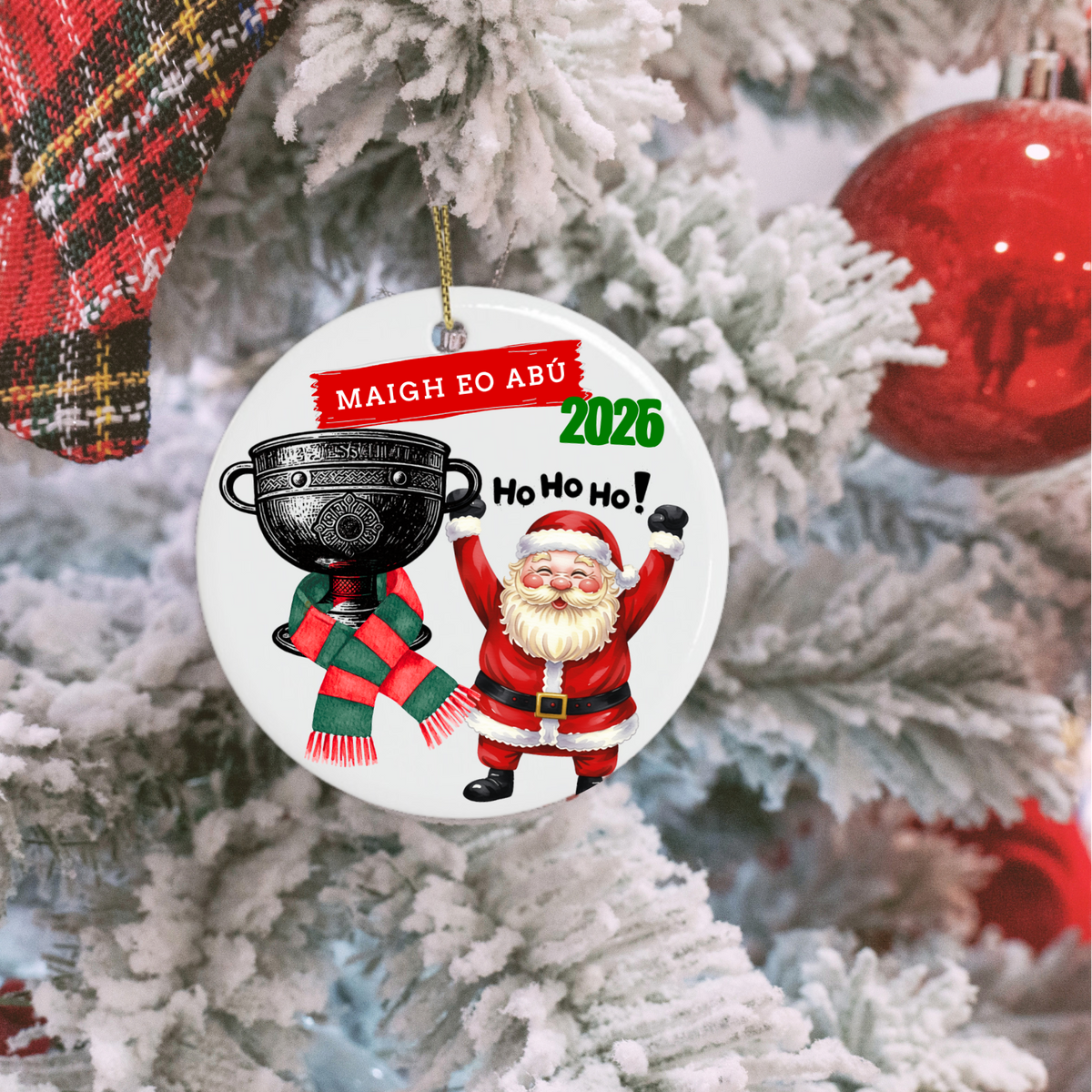 A round white Christmas ornament hanging on a snowy white Christmas tree. The ornament features a printed illustration of Santa Claus holding a black cup, a green and red striped scarf, and festive text reading 'MAIGH EO ABU 2026' and 'Ho Ho Ho!'.