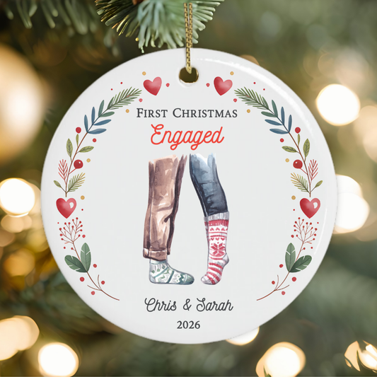 A personalised round ceramic Christmas ornament hanging on a Christmas tree. The white ornament features the text 'FIRST CHRISTMAS Engaged  Chris & Sarah 2026' with an illustration of two legs in holiday clothes, surrounded by a red, green, and gold leafy wreath.