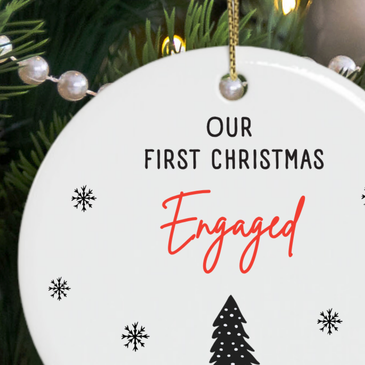 A round white ceramic Christmas ornament gift for engaged couple hanging from a gold string, printed with the words 'OUR FIRST CHRISTMAS Engaged' in black and red script, with small black snowflakes and a tree near the bottom. The ornament is photographed in front of a Christmas tree with lights and pearl garland.