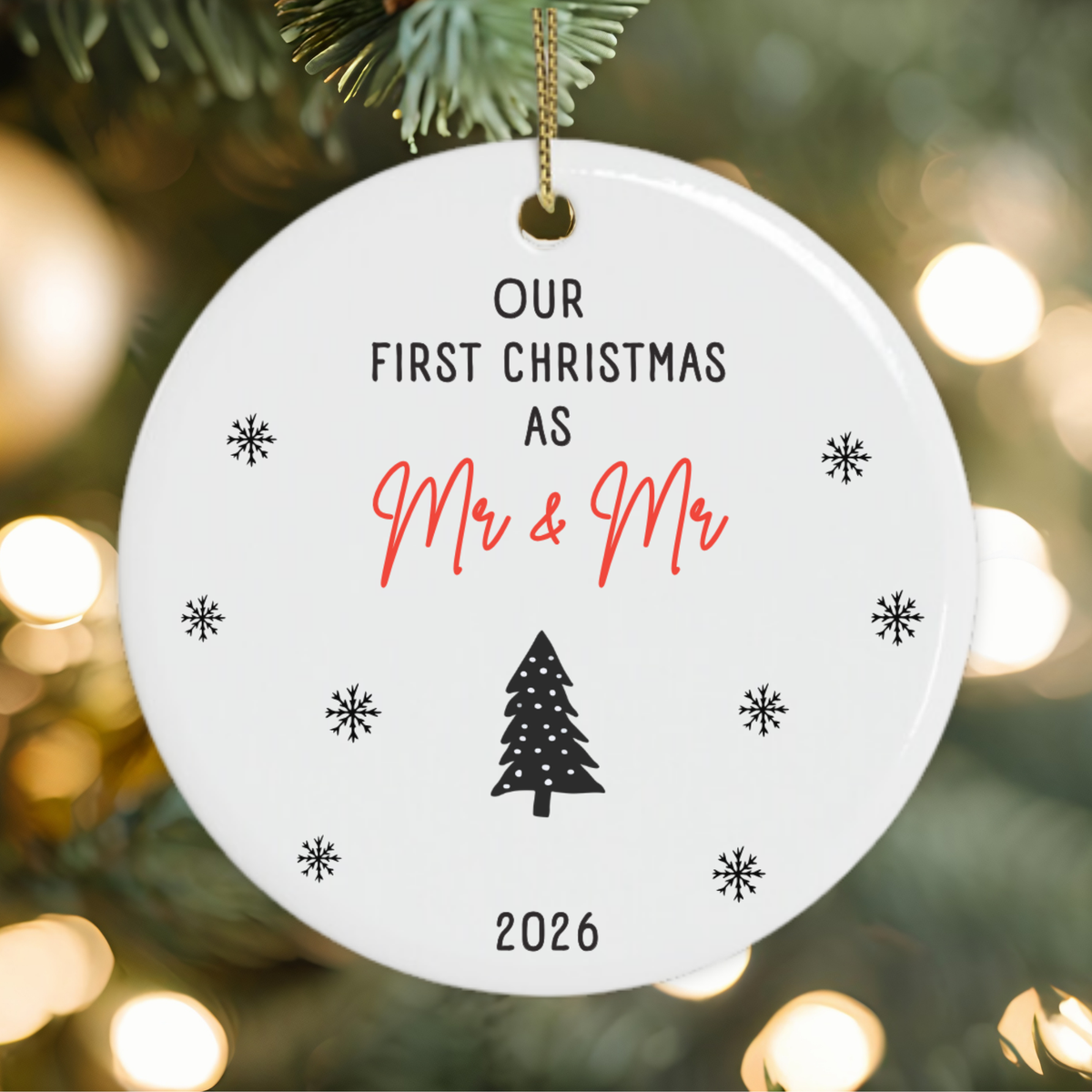 A round white Christmas ornament gift for newlywed gay couple hanging on a decorated tree, printed with the text 'OUR FIRST CHRISTMAS AS Mr & Mr 2026' in black and red script, surrounded by snowflake motifs and a black tree icon.