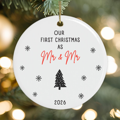 A round white Christmas ornament gift for newlywed gay couple hanging on a decorated tree, printed with the text 'OUR FIRST CHRISTMAS AS Mr & Mr 2026' in black and red script, surrounded by snowflake motifs and a black tree icon.