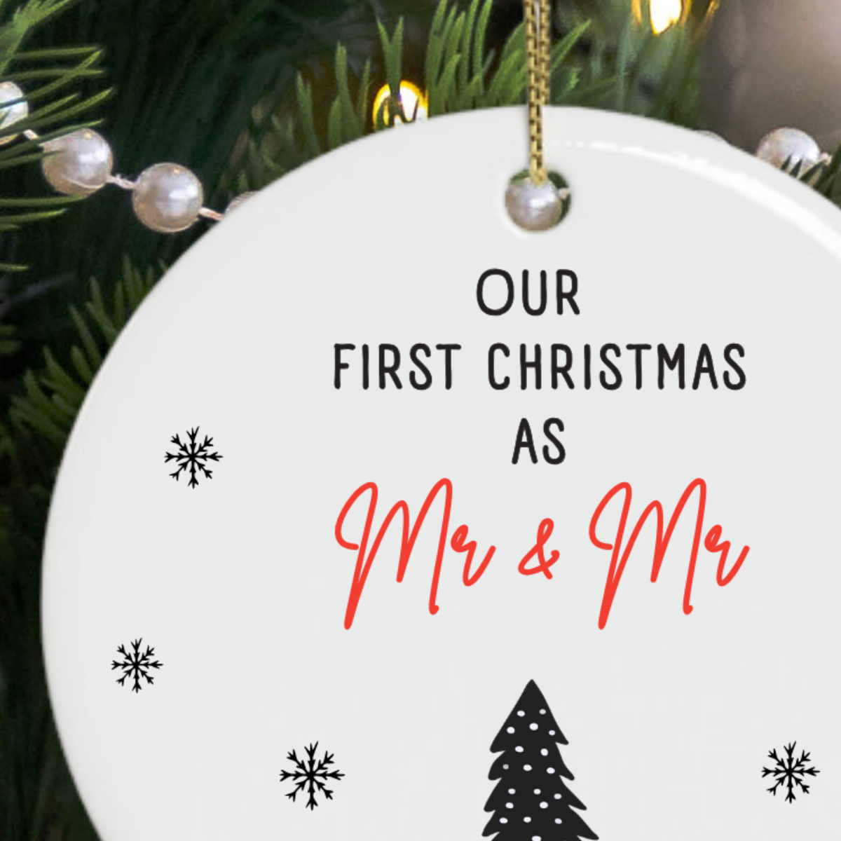 A round white ceramic Christmas ornament gift for newlywed gay couple hanging from a gold string, printed with the text 'OUR FIRST CHRISTMAS AS Mr & Mr' in black and red script, with small black snowflakes and a tree silhouette. 