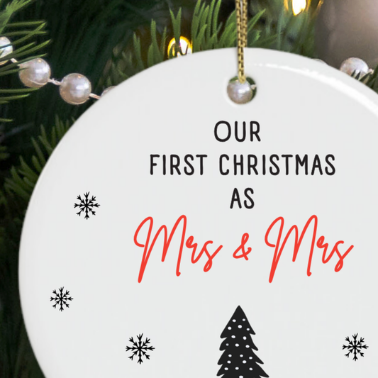 A round white ceramic Christmas ornament gift for newlywed lesbian couple hanging from a gold string, printed with the text 'OUR FIRST CHRISTMAS AS Mrs & Mrs' in black and red script, with small black snowflakes and a tree silhouette. 
