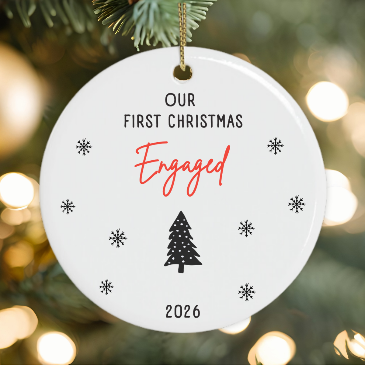 A round white ceramic Christmas ornament hanging from a gold string, printed with the text 'OUR FIRST CHRISTMAS Engaged 2026' along with small black snowflakes and a tree icon, set against a blurred Christmas tree background with warm bokeh lights.