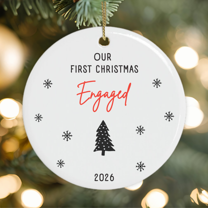 A round white ceramic Christmas ornament hanging from a gold string, printed with the text 'OUR FIRST CHRISTMAS Engaged 2026' along with small black snowflakes and a tree icon, set against a blurred Christmas tree background with warm bokeh lights.