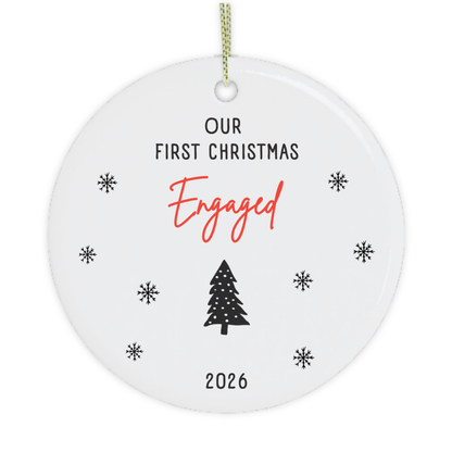 A round white ceramic Christmas ornament with a hanging loop, featuring the text 'OUR FIRST CHRISTMAS Engaged 2026' in black and red lettering, surrounded by small black snowflakes and a black Christmas tree illustration.