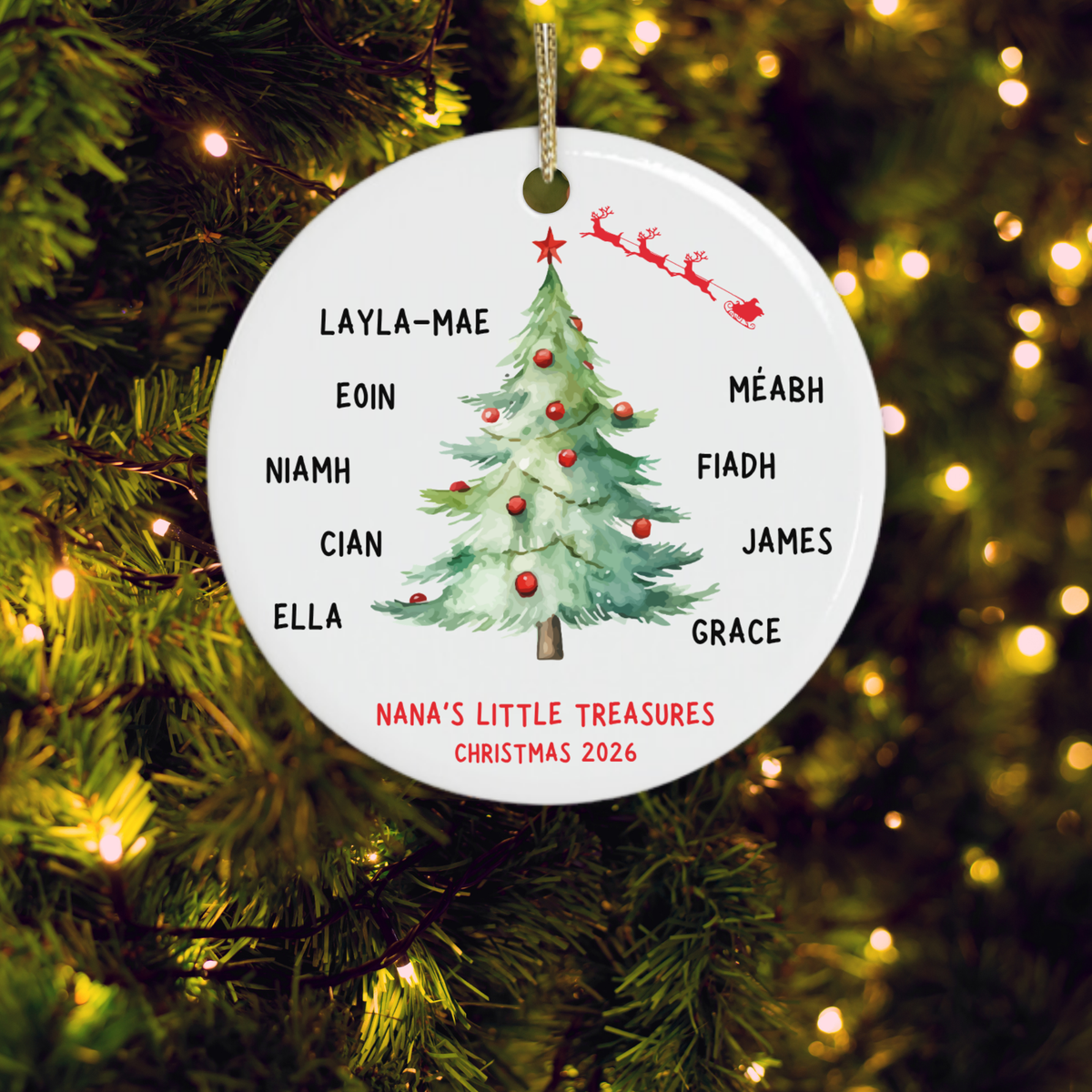 A personalised round ceramic Christmas ornament for grandparents hanging on a lit Christmas tree. The white ornament features a green Christmas tree with red ornaments, Santa in red sleighs, and the names of family members around the border, with the text 'NANA'S LITTLE TREASURES CHRISTMAS 2026' at the bottom.