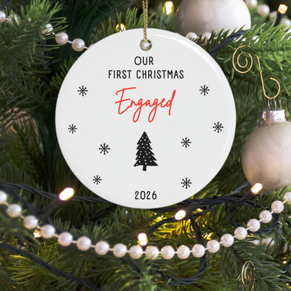 A round white Christmas ornament gift for engaged couple hanging on a decorated tree, printed with the text 'OUR FIRST CHRISTMAS ENGAGED 2026' in black and red script, surrounded by pine branches, pearl beads, and warm string lights.