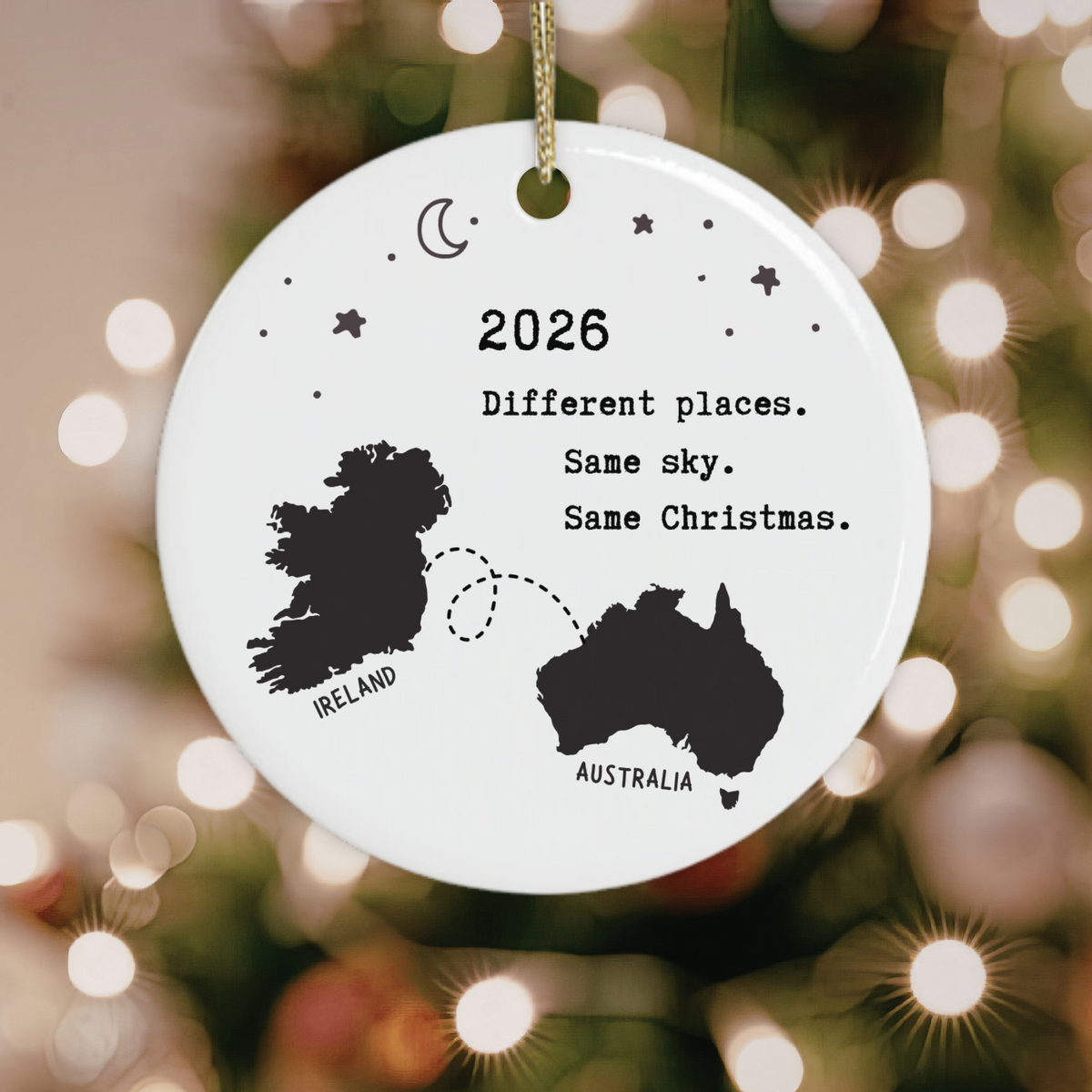 A round white ceramic Christmas ornament hanging from a gold string, printed with black silhouettes of Ireland and Australia, stars, a crescent moon, and the text '2026 Different places. Same sky.