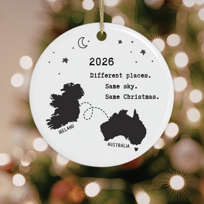 A round white ceramic Christmas ornament hanging from a gold string, printed with black silhouettes of Ireland and Australia, stars, a crescent moon, and the text '2026 Different places. Same sky.