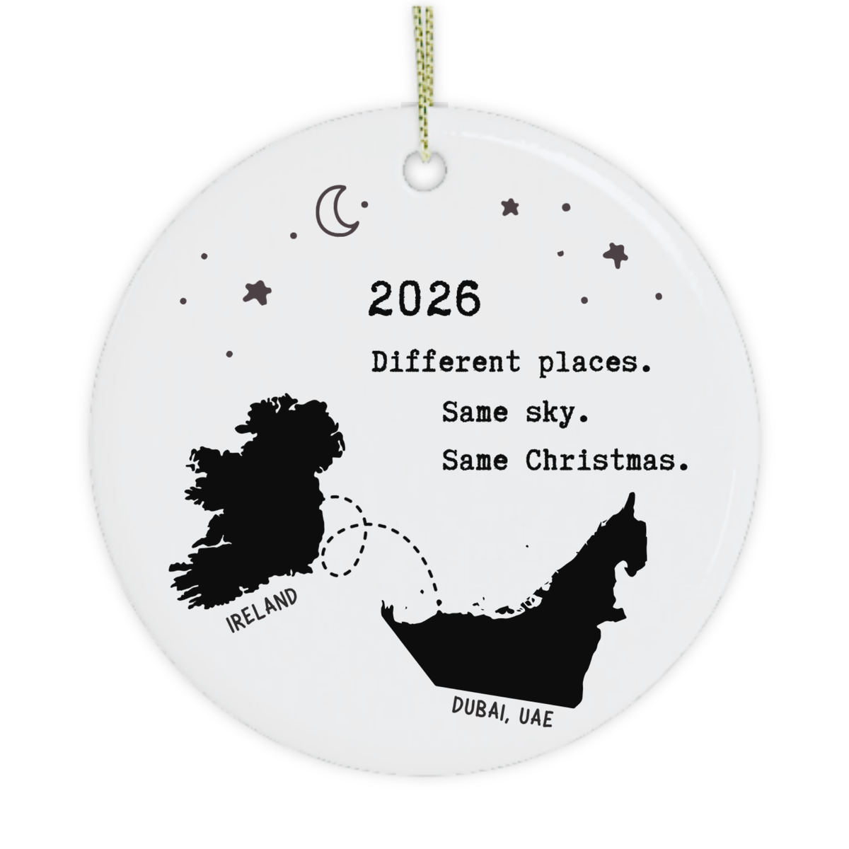 A round white ceramic Christmas ornament with a gold hanging string, featuring black printed graphics of Ireland and the United Arab Emirates with the text '2026 Different places. Same sky.