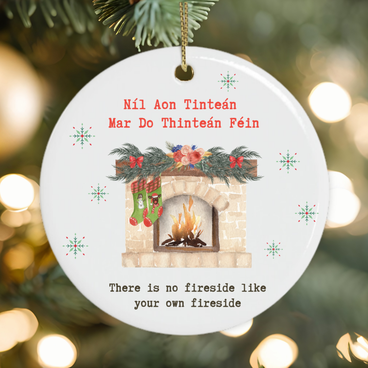 A round white ceramic Christmas ornament hanging from a gold string on a pine tree branch. The ornament features a printed illustration of a cozy fireplace with stockings, a garland, and snowflakes, along with the Irish text 'Éil Aon Tinteán Óar Do Tinteán Féin' and the English phrase 'There is no fireside like your own fireside' against a blurred holiday background with warm bokeh lights.