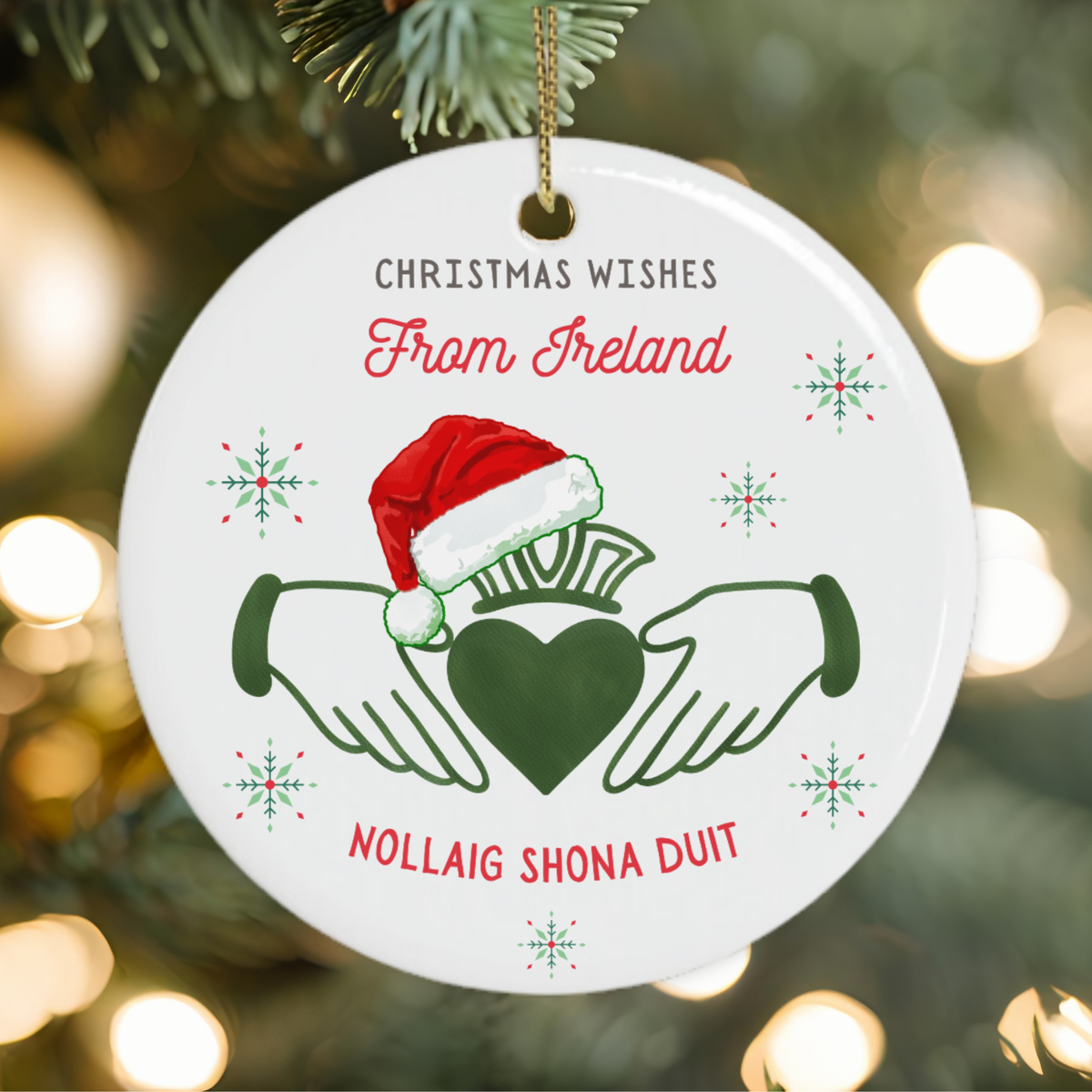 A round white Christmas ornament hanging from a tree branch, printed with 'CHRISTMAS WISHES From Ireland' and 'NOLLAIG SHONA DUIT' around a green heart and hands design with a red Santa hat, against a blurred christmas tree background with warm lights.