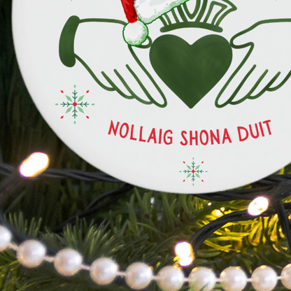 A close-up of a round white Christmas ornament hanging on a decorated Christmas tree. The ornament features a green Claddagh symbol with a crown and a green heart, red text reading 'NOLLAIG SHONA DUIT', and small red and green snowflakes.