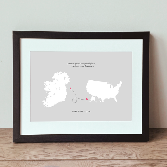 Framed travel-style art print showing white map silhouettes of Ireland and the United States with a small pink heart connecting them, displayed on a wooden surface against a light wall.