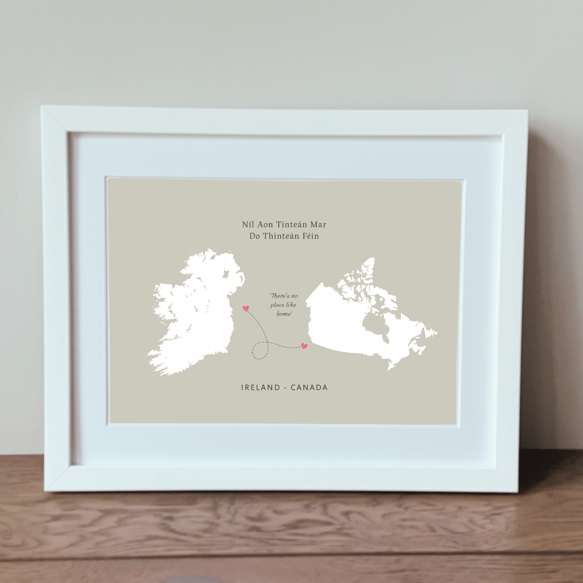 A framed travel-style map print showing Ireland and Canada with a connecting line and small pink heart accents, displayed in a white frame on a wooden surface against a light neutral wall.