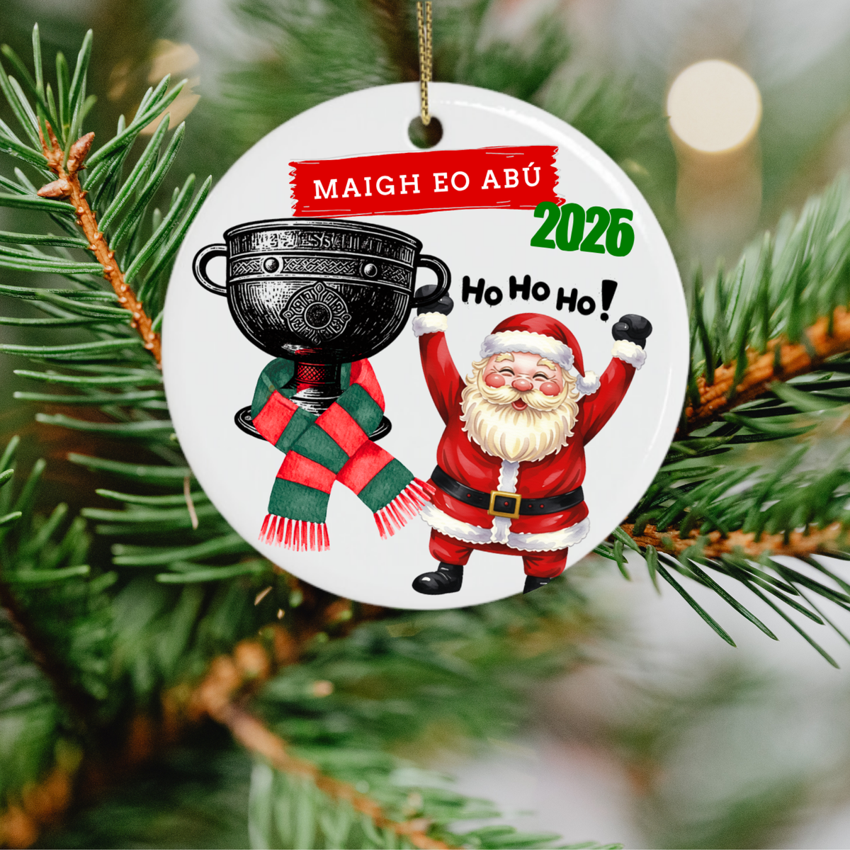 A round white ceramic Christmas ornament hanging from a gold string on a pine tree branch. The ornament features a printed illustration of Santa Claus holding a large black trophy and wearing a red-and-green scarf, with the text “MAIGH EO ABÚ»” in red, “2026” in green, and “Ho Ho Ho!” in black.