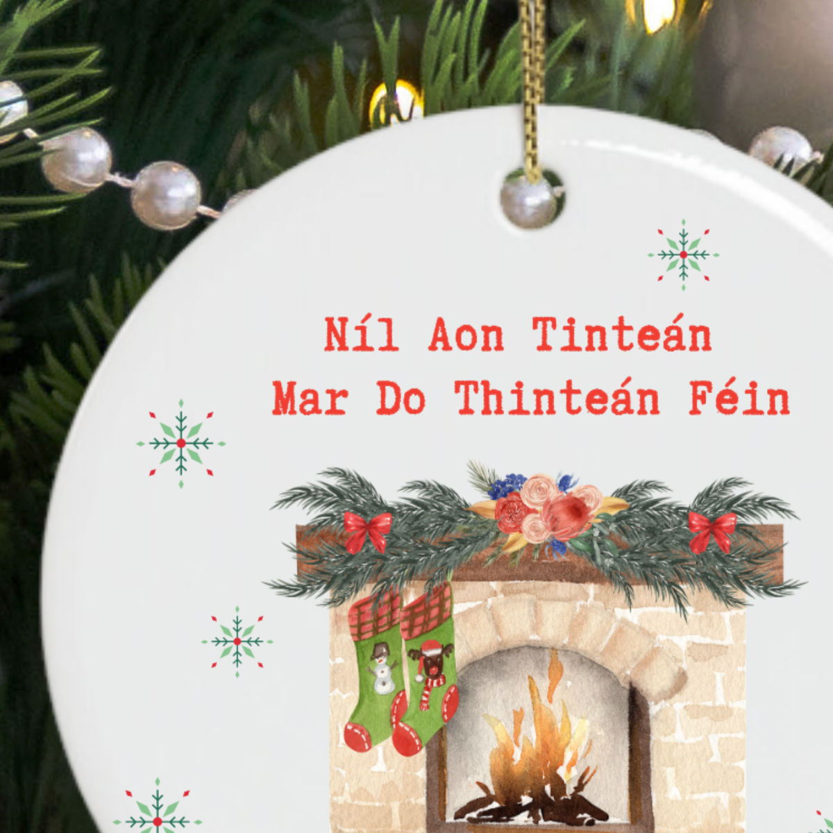 A round white ceramic Christmas ornament hanging from a gold string, featuring red Irish text reading 'Níl Aon Tinteán Mar Do Thinteán Féin' above an illustration of a fireplace with green stockings, evergreen garland, and snowflake motifs, set against a background of pine branches and lights.