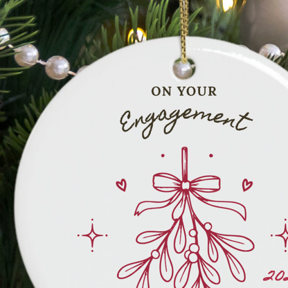A round white ceramic personalised Christmas ornament hanging from a gold string, printed with the words 'ON YOUR Engagement' and a red mistletoe illustration, shown against a Christmas tree background.
