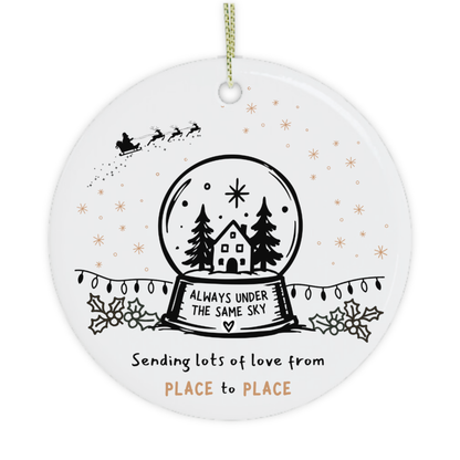 A round white ceramic Christmas ornament with a hanging loop, featuring a black line-art snow globe with a house and trees, Santa in the sky, and decorative text reading 'ALWAYS UNDER THE SAME SKY' and 'Sending lots of love from PLACE to PLACE' on a white background.