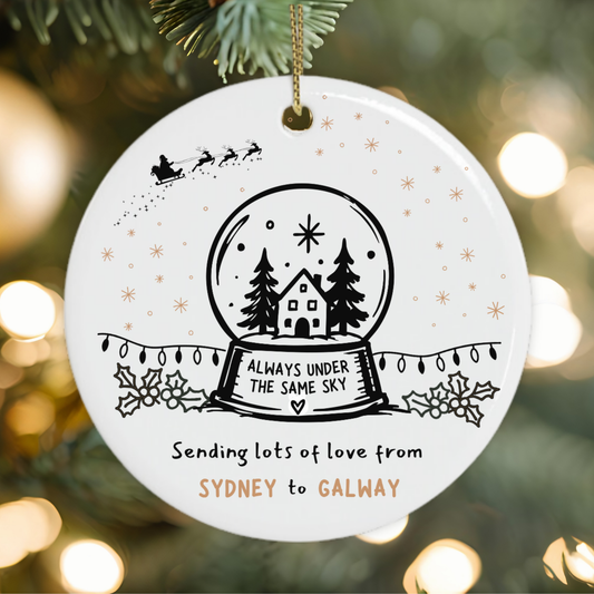 A round white ceramic Christmas ornament hanging on a tree branch, featuring a black snow globe illustration with a house and trees, gold star accents, and personalized text reading 'Sending lots of love from SYDNEY to GALWAY'.