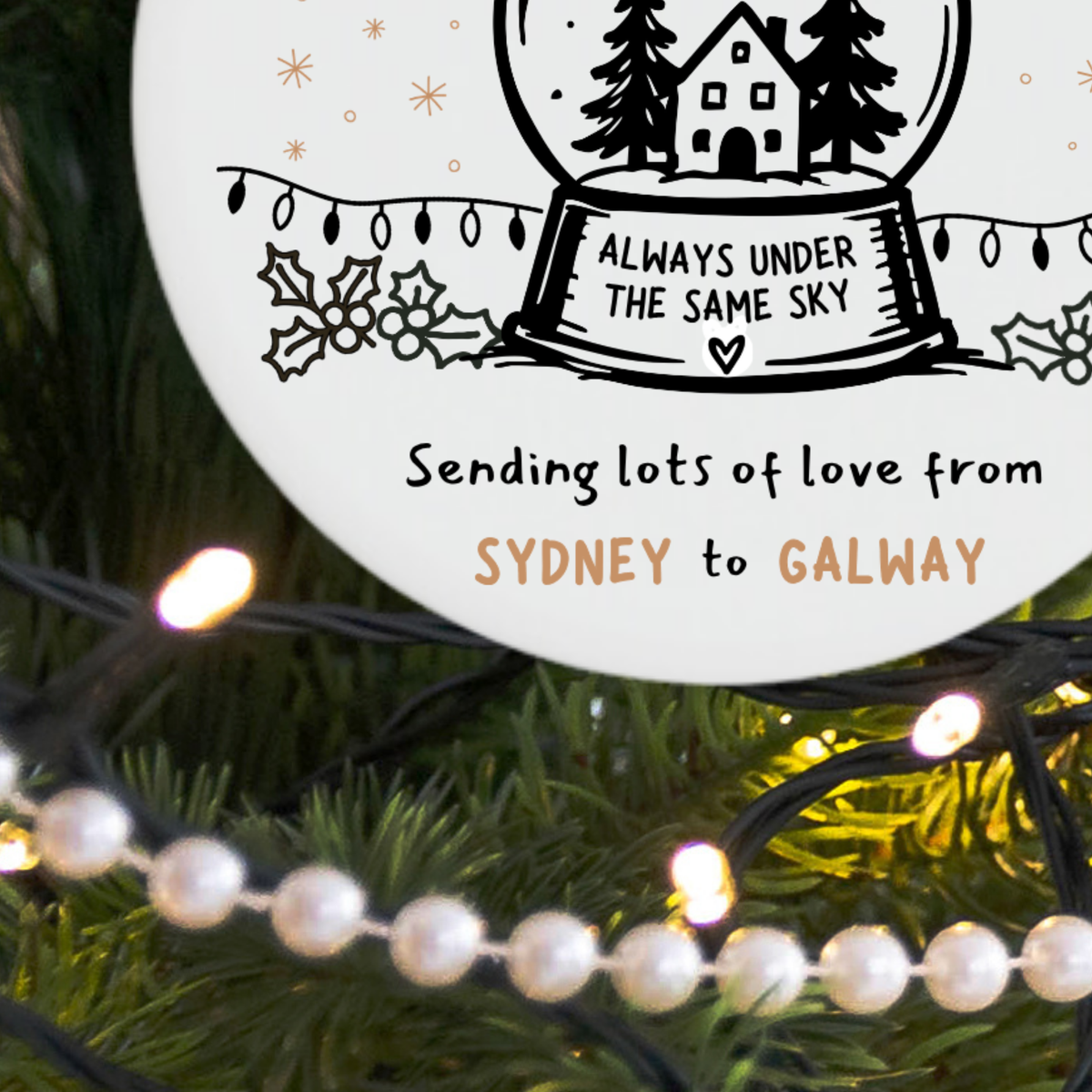 A round white holiday ornament or plaque with a snow globe illustration and the text 'Always Under the Same Sky' and 'Sending lots of love from Sydney to Galway,' displayed on a green tree with warm white string lights.