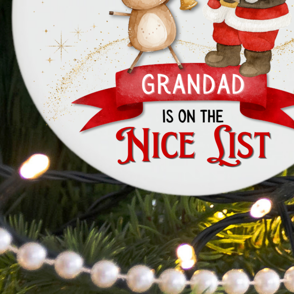 A round white Christmas ornament with festive illustrations of a reindeer and Santa Claus, featuring the text 'GRANDAD IS ON THE NICE LIST' in red and black. The ornament is shown hanging in front of a Christmas tree with warm string lights and pearl beads.