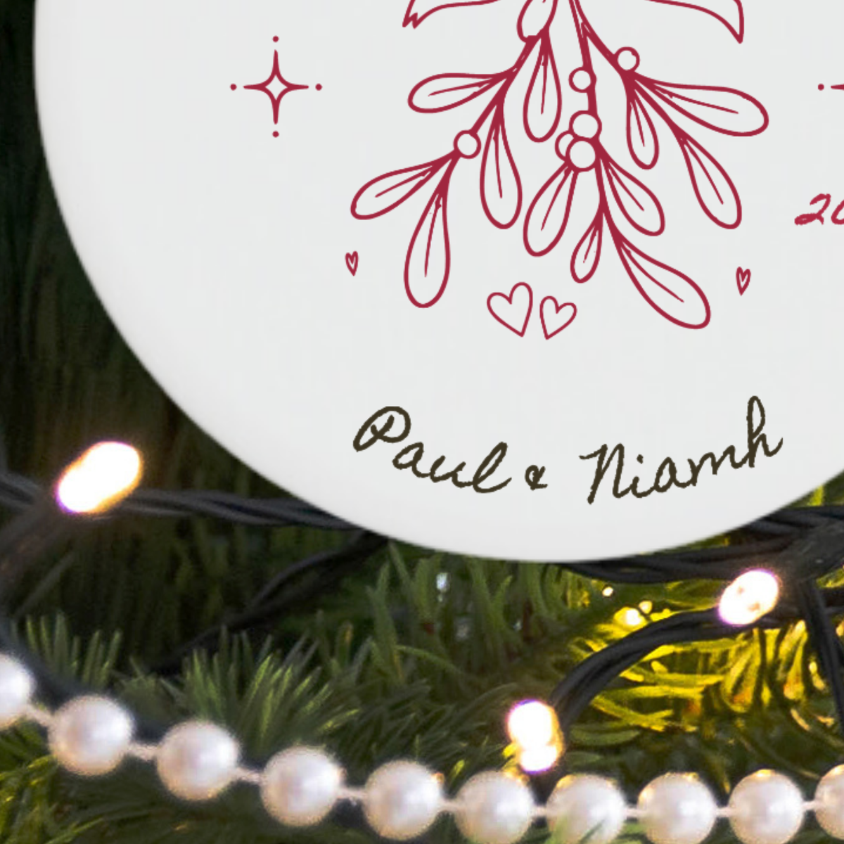 A close-up of a round white personalised Christmas ornament with red mistletoe illustrations and the sample names 'Paul & Niamh' in black script, shown hanging among green pine branches and warm white string lights.