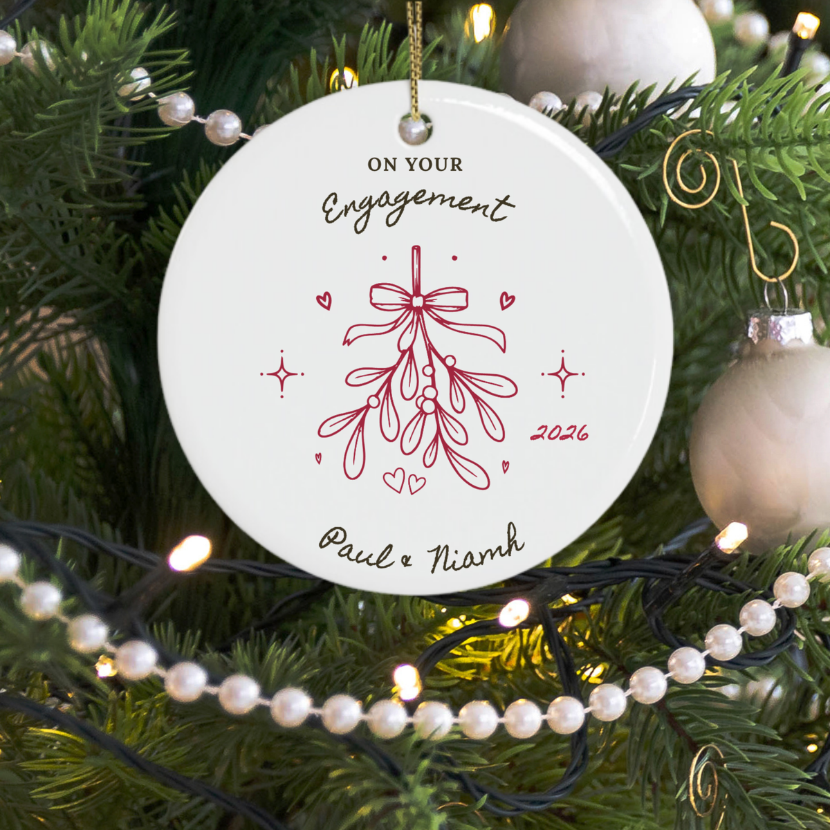 A round white personalised ceramic Christmas ornament hanging on a decorated Christmas tree. The ornament features a red mistletoe illustration with the words 'ON YOUR Engagement 2026' and personalised names 'Paul & Niamh' in black script.
