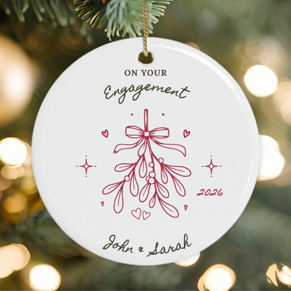 A round white ceramic personalised Christmas ornament hanging from a gold string, printed with the words 'ON YOUR Engagement' and a red mistletoe illustration, shown against a Christmas tree background.
