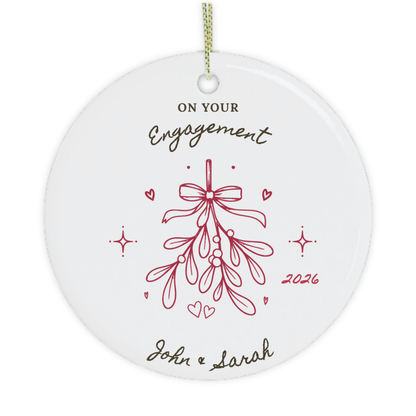 A round white ceramic personalised Christmas ornament with a hanging loop, featuring the text 'on your engagement 2026' in black and red lettering, with a mistletoe illustration.